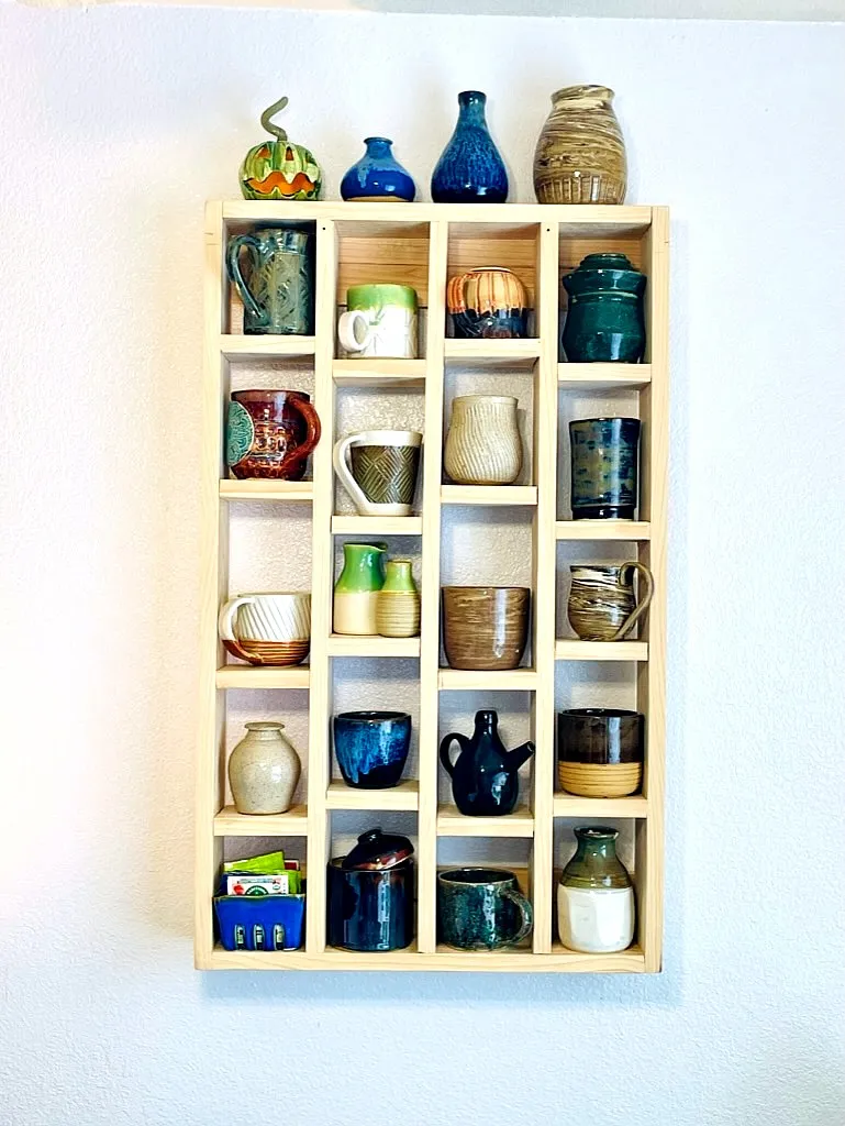 A wall shelf filled with handmade mugs, vases, pitchers, and lidded jars in varied glazes